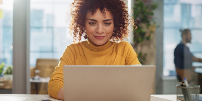 Photo of woman of color facing the camera and typing on a laptop